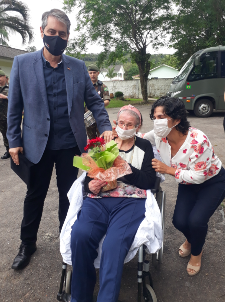 Idosa em cadeira de rodas segura um buquê de flores vermelhas, acompanhada por um homem de terno azul escuro à esquerda e uma mulher de blusa branca com flores vermelhas à direita, ambos usando máscaras. Ao fundo, aparecem soldados, uma árvore, casas e um micro-ônibus com bandeira do Brasil.
