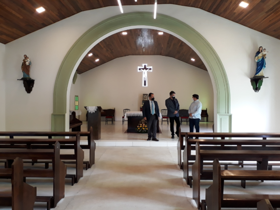 Três homens de máscara conversam próximos ao altar de uma capela simples e bem iluminada, com bancos de madeira vazios em primeiro plano. A capela possui teto de madeira, imagens de santos nas paredes laterais e uma cruz iluminada ao fundo, acima do altar.