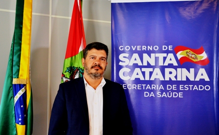 Homem de terno escuro e camisa branca posa em frente a um banner do Governo de Santa Catarina, Secretaria de Estado da Saúde.