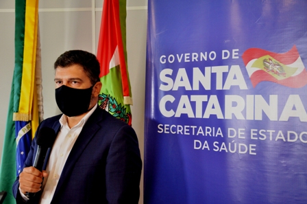 Homem usando terno escuro, camisa branca e máscara preta segura um microfone em frente a um banner do Governo de Santa Catarina, Secretaria de Estado da Saúde.