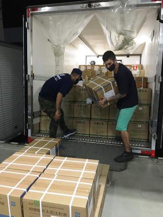 Dois homens vestindo roupas pretas descarregam caixas marrons da caçamba de um caminhão durante a noite.