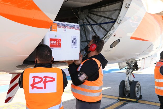 Homem vestindo colete laranja de identificação descarrega uma caixa térmica branca da carga de um avião, em uma operação de recebimento de vacinas contra a COVID-19. A cena ocorre em um aeroporto de Santa Catarina (2021).
