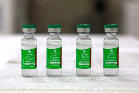 Quatro frascos da vacina Covishield (ChAdOx1 nCoV-19), desenvolvida pela AstraZeneca em parceria com a Universidade de Oxford, estão posicionados lado a lado sobre uma superfície branca. Cada frasco tem tampa verde e rótulo verde com informações sobre o imunizante.