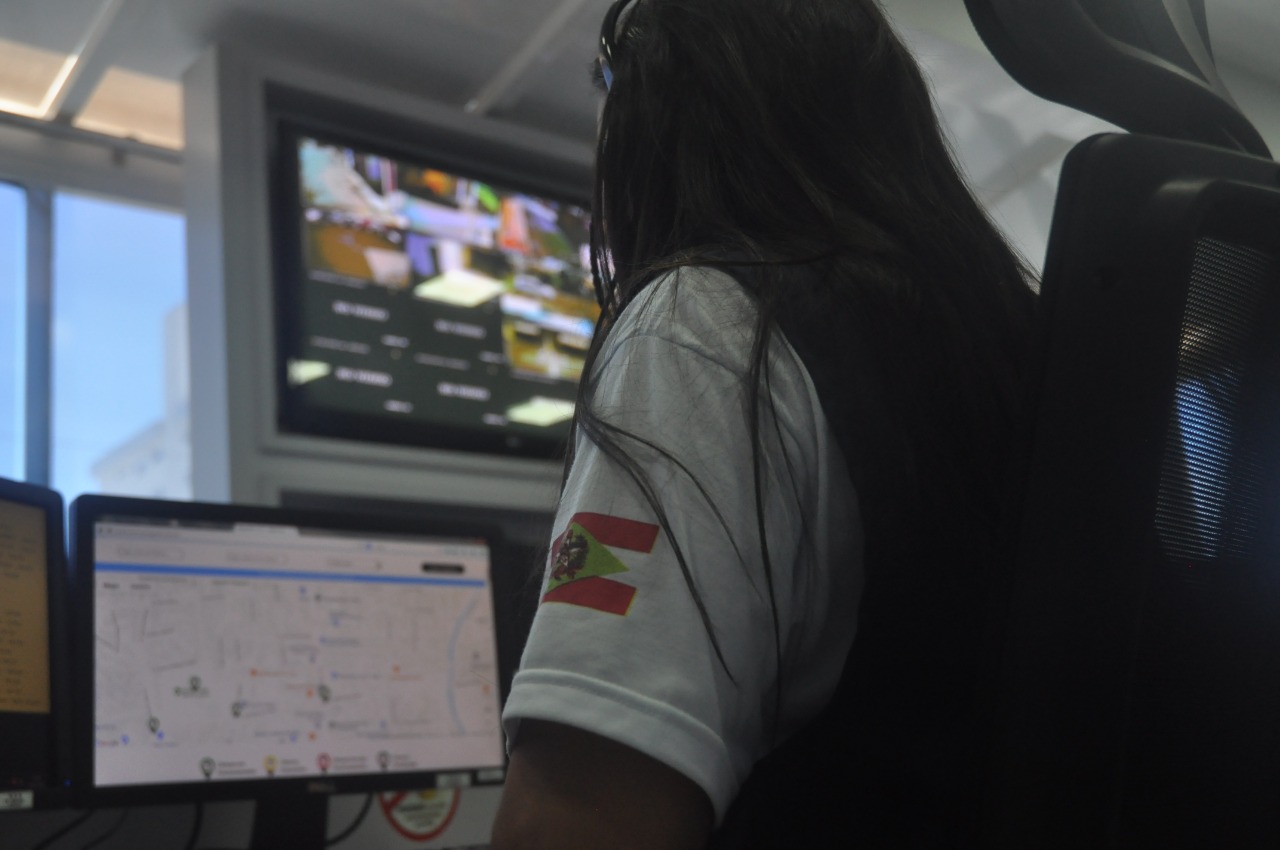 Pessoa de costas usando fone de ouvido e camiseta branca com a bandeira de Santa Catarina no braço. Ela está em uma central de monitoramento, olhando para uma tela de computador com um mapa exibido. Ao fundo, há um monitor grande na parede com imagens de câmeras de vigilância.