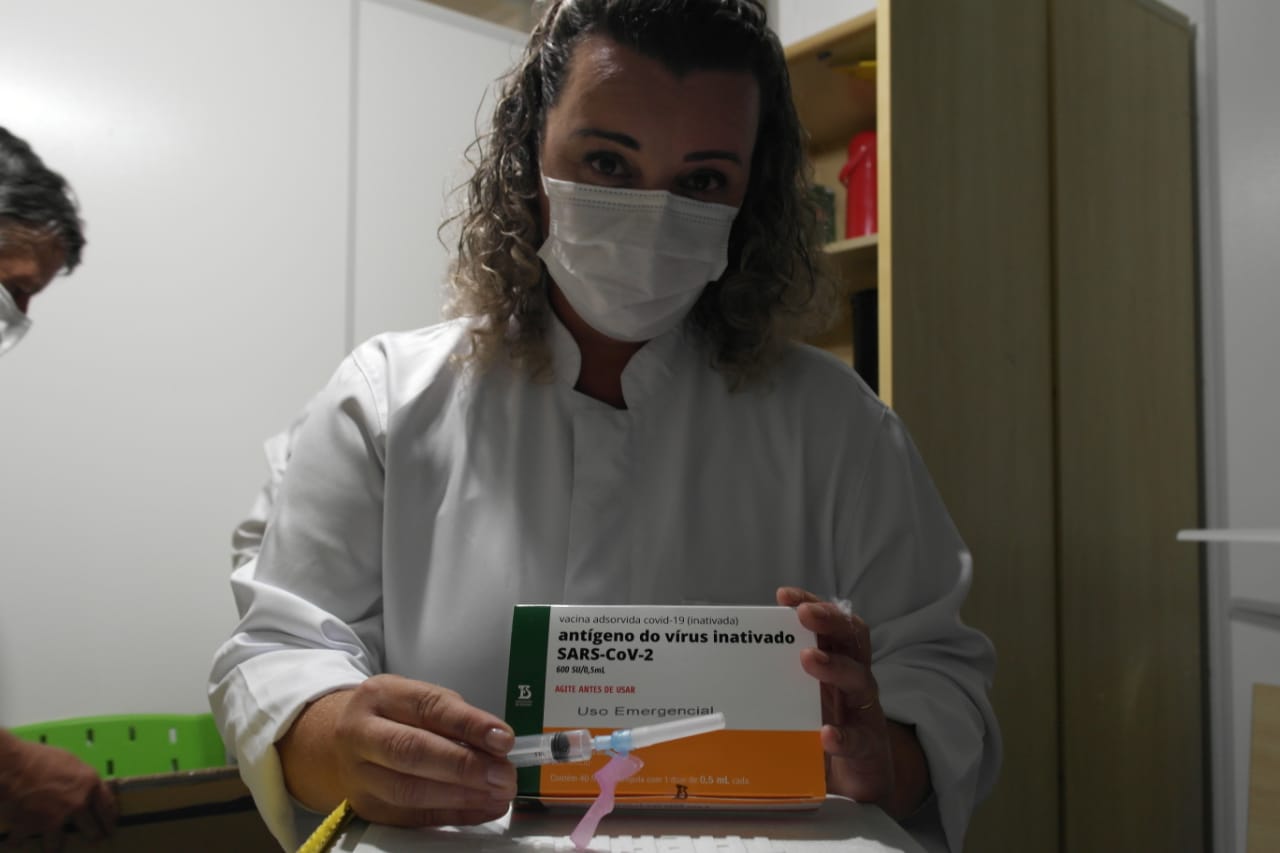 Profissional de saúde com jaleco branco e máscara segura uma caixa da vacina contra a COVID-19 com os dizeres antígeno do vírus inativado SARS-CoV-2 e Uso Emergencial. Ela também segura uma seringa pronta para aplicação. Ao fundo, há armários de madeira e outro profissional parcialmente visível.