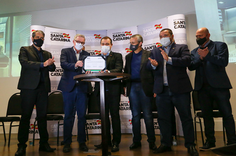 Evento oficial com seis homens em pé sobre um palco, todos de máscara, em frente a painéis com a marca do Governo de Santa Catarina. Ao centro, duas autoridades seguram e apresentam um documento. Os demais aplaudem a ação. A imagem transmite um momento de entrega ou assinatura de documento oficial.