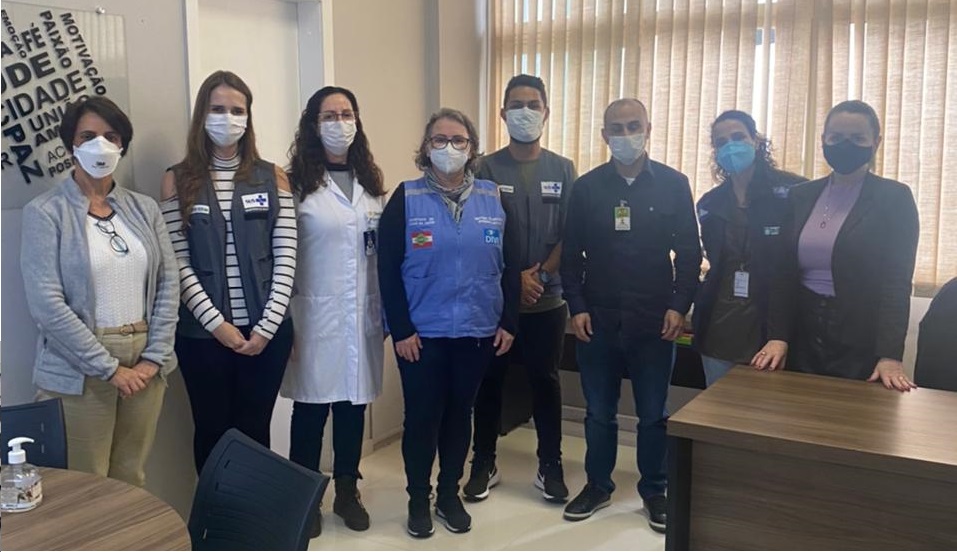 Grupo de nove profissionais da saúde posa para foto em uma sala iluminada por luz natural, com persianas ao fundo. Todos usam máscaras de proteção facial e crachás. Entre eles, há pessoas com jaleco branco, coletes identificadores e roupas sociais. À esquerda, uma mesa redonda com cadeiras e um frasco de álcool em gel. A parede exibe uma arte com palavras motivacionais.