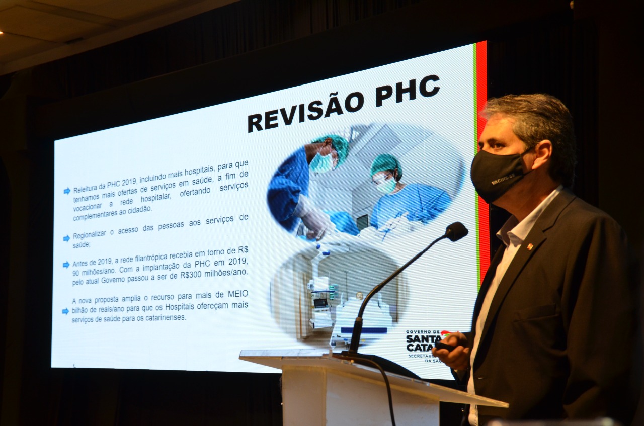 Imagem de apresentação sobre a Revisão PHC – Governo de Santa Catarina:  À direita, um homem de terno escuro e máscara preta com a palavra "VACINE-SE" fala ao microfone em um púlpito. Ao fundo, há um telão exibindo um slide com o título "REVISÃO PHC". O conteúdo da apresentação inclui:  Releitura da PHC 2019, com inclusão de mais hospitais para ampliar a oferta de serviços em saúde e oferecer serviços complementares ao cidadão.  Regionalização do acesso da população aos serviços de saúde.  Antes de 2019, a rede filantrópica recebia cerca de R$ 90 milhões por ano. Após a implantação da PHC em 2019, esse valor passou para R$ 300 milhões por ano.  A nova proposta amplia o recurso para mais de meio bilhão de reais ao ano.  O slide contém também a logomarca do Governo de Santa Catarina e uma imagem circular com dois profissionais da saúde de avental e touca cirúrgica realizando um procedimento hospitalar.