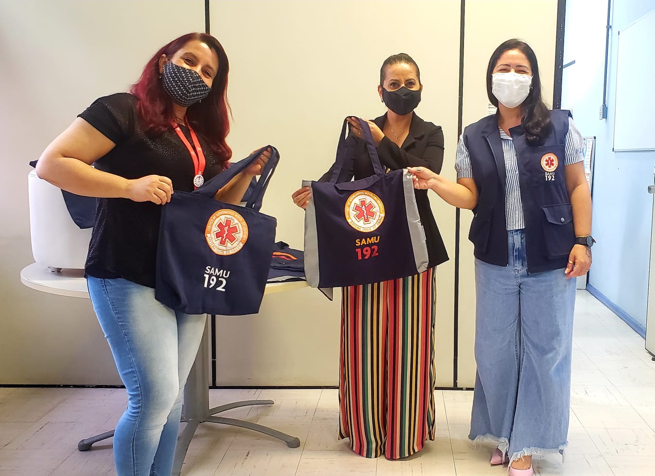 'Três mulheres posam sorrindo, usando máscaras de proteção facial, enquanto seguram bolsas do SAMU com o número 192 estampado. As bolsas são azul-escuras com o símbolo da emergência médica em destaque. À esquerda, uma mulher usa blusa preta e jeans claro; ao centro, outra veste blazer preto e calça de listras coloridas; à direita, a terceira usa colete do SAMU sobre camisa listrada e calça jeans larga. Elas estão em um ambiente interno, com piso claro e divisórias ao fundo.