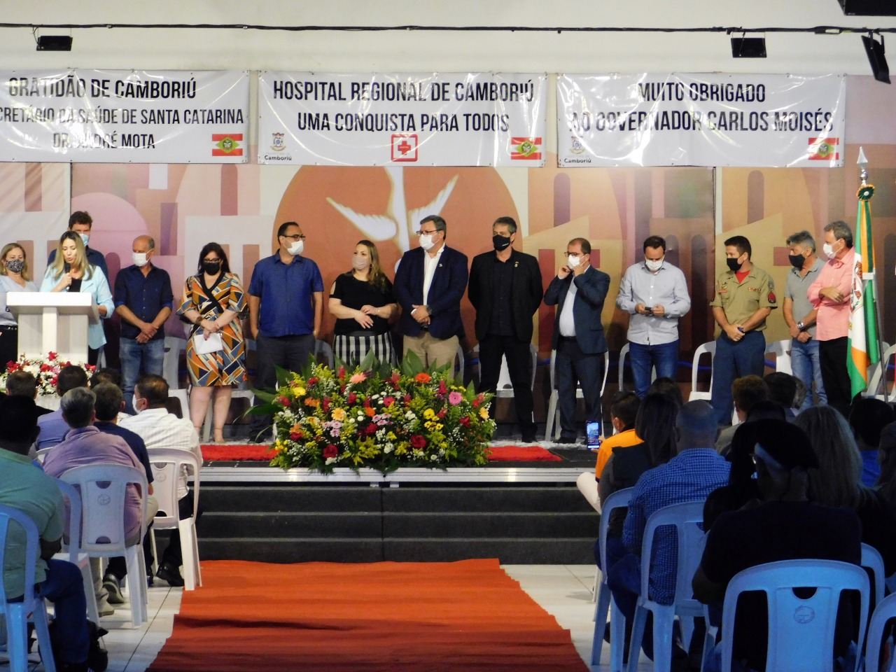 Evento oficial com diversas autoridades em um palco decorado com flores e banners. Um dos cartazes diz "Hospital Regional de Camboriú — Uma conquista para todos". Pessoas estão usando máscaras, e há público sentado em cadeiras brancas assistindo à cerimônia. Ao fundo, bandeiras de Santa Catarina e do município estão posicionadas.
