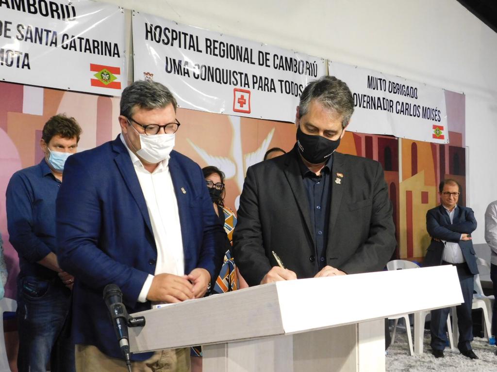 Duas autoridades assinando documento em cerimônia oficial no palco. Ambos usam máscara, sendo que um deles está vestindo terno azul e o outro, terno preto. Ao fundo, há banners com os dizeres “Hospital Regional de Camboriú – Uma conquista para todos” e agradecimentos ao governador Carlos Moisés. Outras pessoas, também de máscara, observam a cena.