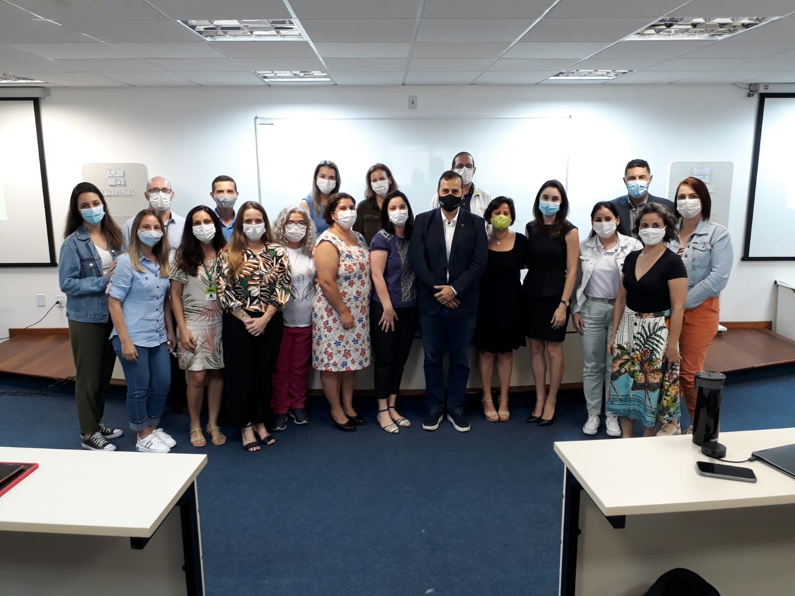 A imagem mostra um grupo de aproximadamente 22 pessoas, reunidas em uma sala de aula ou auditório. Todos estão usando máscaras, indicando cuidados com a saúde, provavelmente relacionados à pandemia de COVID-19. O ambiente parece ser institucional, com projetores e quadro branco ao fundo, e o logotipo da UDESC (Universidade do Estado de Santa Catarina) visível na parede à esquerda. O grupo está posicionado de forma organizada, como em um registro de evento, capacitação ou encerramento de curso.