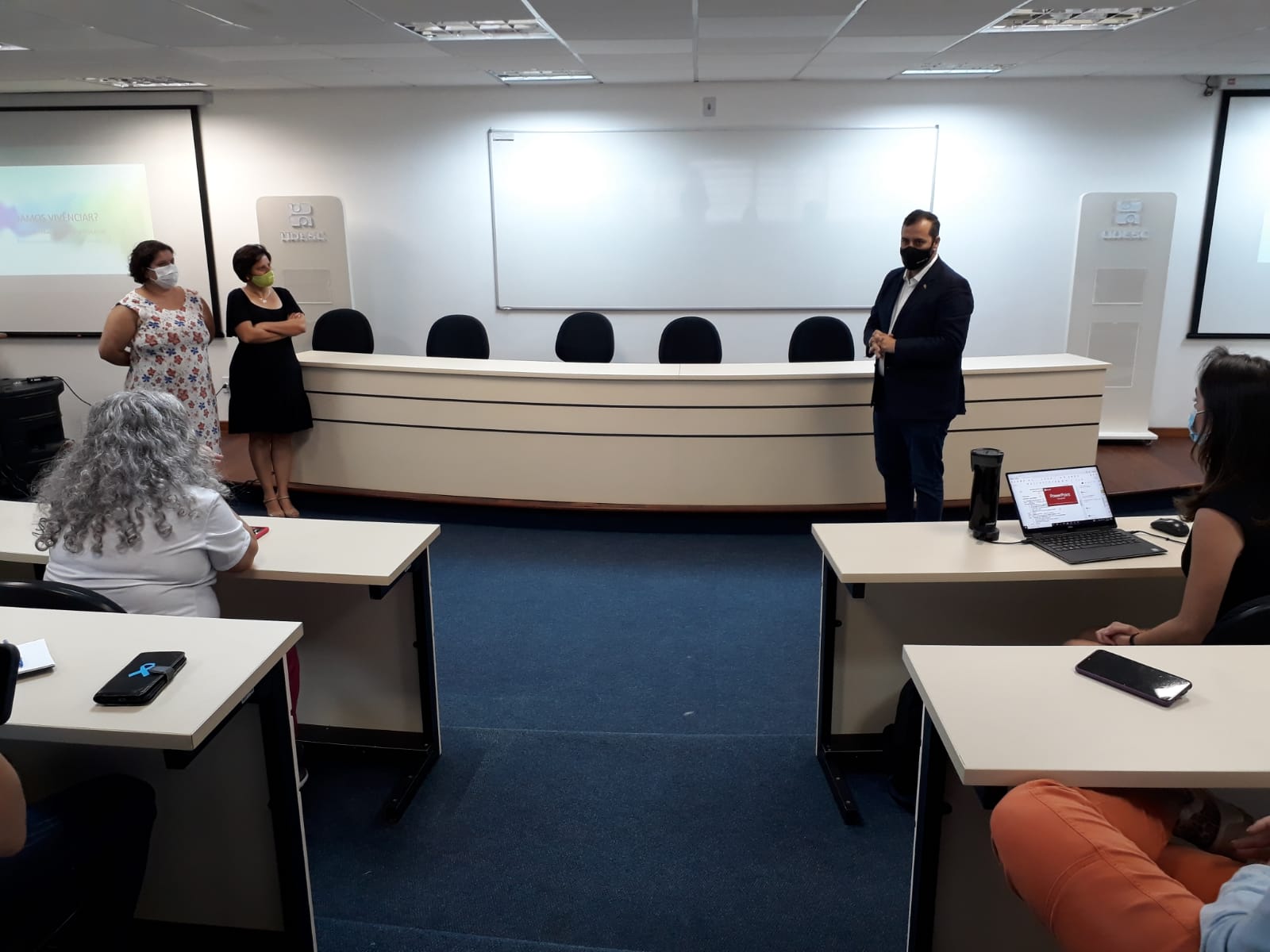 Essa imagem mostra um momento de apresentação ou discurso em uma sala de aula ou auditório institucional. Três pessoas estão em pé à frente da sala, uma delas aparenta estar discursando, enquanto outras assistem sentadas. Todas usam máscara, respeitando protocolos sanitários. O logotipo da UDESC aparece novamente ao fundo, indicando que o evento está sendo realizado na Universidade do Estado de Santa Catarina.