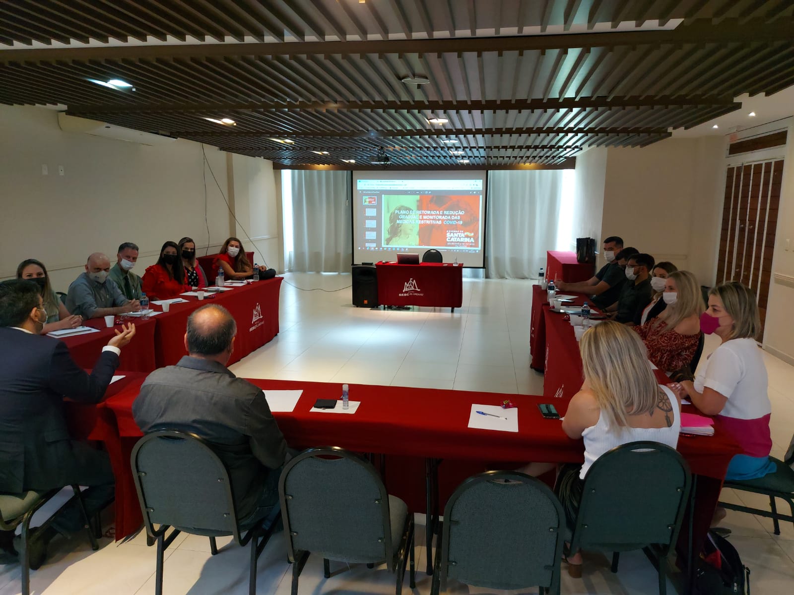 Reunião com cerca de 20 pessoas sentadas em formato de U em uma sala de conferência com mesas cobertas por toalhas vermelhas. Ao fundo, uma apresentação é projetada com o título “PLANO ESTRATÉGICO REDUÇÃO DA MORTALIDADE MATERNA DAS MULHERES CATARINENSES”. Todos os participantes utilizam máscara, demonstrando cumprimento dos protocolos sanitários. A sala possui iluminação natural e teto com ripado de madeira.