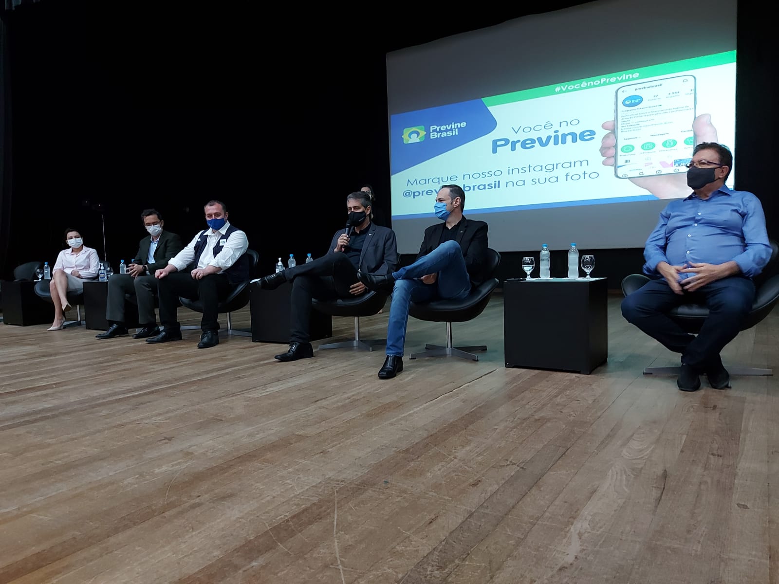 Seis pessoas sentadas em poltronas pretas sobre um palco de madeira, participando de um painel ou debate. Todos usam máscara facial. Um dos participantes segura um microfone enquanto fala. Ao fundo, um telão exibe uma apresentação com o logotipo do programa 'Previne Brasil' e a frase 'Você no Previne - Marque nosso instagram @previnebrasil na sua foto'. Há garrafas de água e taças sobre pequenos móveis entre os participantes.