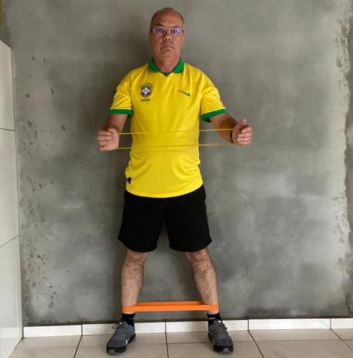 Homem de meia-idade em pé diante de uma parede cinza, usando camisa amarela da seleção brasileira, shorts pretos e tênis. Ele segura um elástico de resistência com os braços estendidos à frente do corpo e outro elástico laranja preso nas pernas, na altura dos tornozelos, indicando que está realizando um exercício físico de resistência.