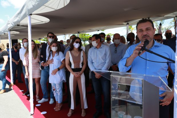 Homem de camisa azul fala ao microfone em um púlpito transparente durante um evento ao ar livre sob uma tenda. Atrás dele, há um grupo de pessoas, muitas usando máscara, incluindo homens e mulheres com roupas sociais. O evento ocorre sobre um tapete vermelho e parece ser uma cerimônia oficial ou solenidade pública.