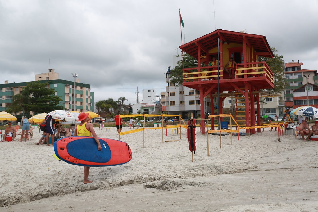 Praia com céu nublado e movimentação de banhistas. Em destaque, um salva-vidas carrega uma prancha de resgate em direção ao mar, vestindo uniforme amarelo e vermelho. Ao fundo, um posto de observação elevado pintado nas mesmas cores abriga outros guarda-vidas. Há guarda-sóis, pessoas sentadas e praticando esportes na areia, além de prédios residenciais ao redor.
