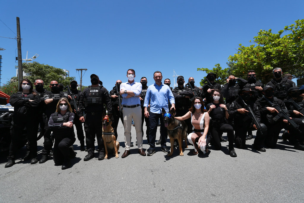 Grupo de policiais fortemente armados posa para foto ao ar livre sob céu azul. Ao centro, dois homens de camisa social e duas mulheres, todos usando máscara, estão acompanhados de cães policiais. As mulheres estão agachadas ao lado dos cães, que usam equipamentos de proteção. A maioria dos policiais veste uniforme preto tático e colete à prova de balas. A imagem transmite uma ocasião oficial ou apresentação da corporação.