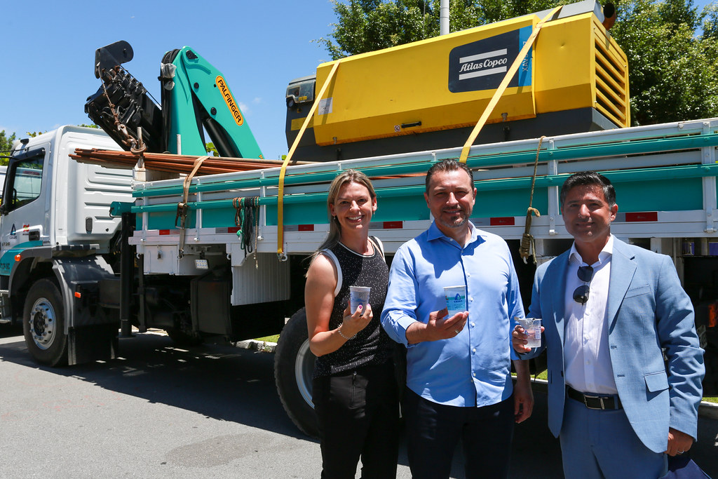 Três pessoas sorridentes seguram copos de água posando em frente a um caminhão equipado com maquinário da marca Atlas Copco e uma grua Palfinger. O grupo é composto por uma mulher e dois homens, todos vestidos com roupas sociais. A cena ocorre em via pública sob céu azul, indicando provável evento de entrega de equipamento ou ação ligada a serviços públicos de abastecimento ou perfuração de poços.