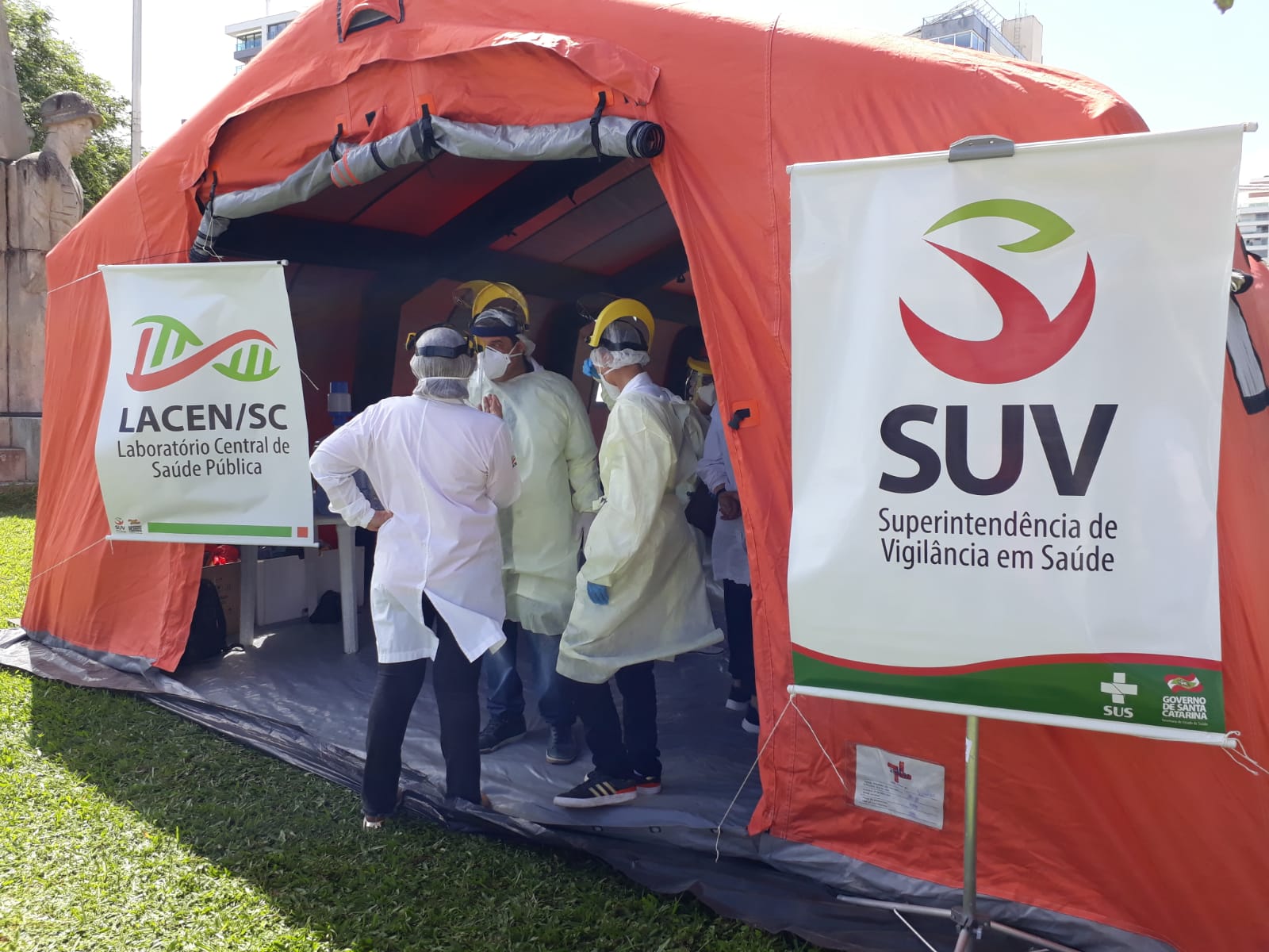 Profissionais de saúde com equipamentos de proteção individual estão reunidos na entrada de uma tenda inflável laranja montada em área externa gramada. Dois banners identificam as instituições envolvidas: LACEN/SC (Laboratório Central de Saúde Pública) e SUV (Superintendência de Vigilância em Saúde). A ação é promovida pelo Governo de Santa Catarina e o SUS, sugerindo atividade relacionada à testagem ou vigilância epidemiológica.
