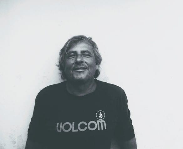 Homem de meia-idade sorri levemente em frente a uma parede branca, vestindo uma camiseta escura com o logo da marca Volcom. A imagem é em preto e branco.