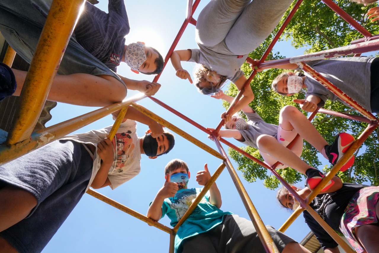 A imagem mostra um grupo de crianças brincando em uma estrutura metálica de escalada em um parque ou escola, vista de baixo para cima. Elas usam máscaras faciais, indicando que a foto foi tirada durante o período da pandemia de COVID-19. A cena transmite um clima alegre e ensolarado, com céu azul e árvores ao fundo, e destaca a interação social e a diversão infantil mesmo em tempos de precaução sanitária.