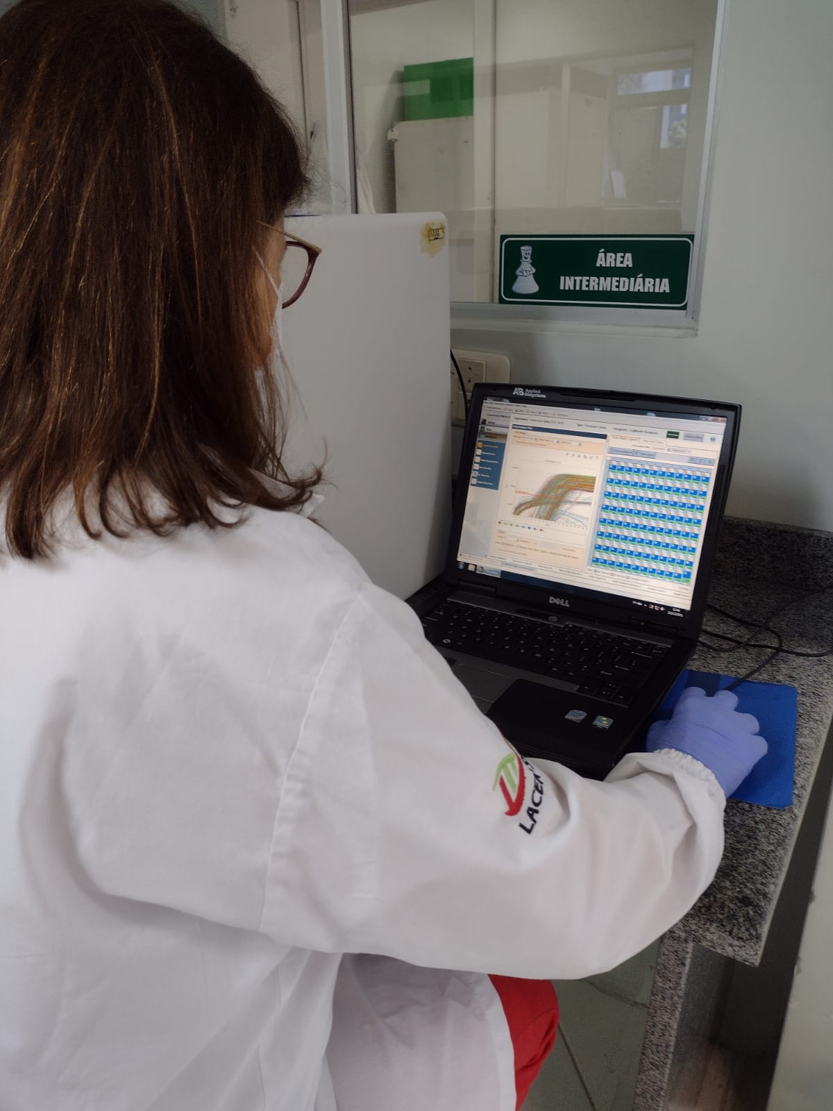 Profissional de laboratório usando jaleco e luvas analisa gráficos em um notebook dentro de uma área com a placa 'Área Intermediária'.