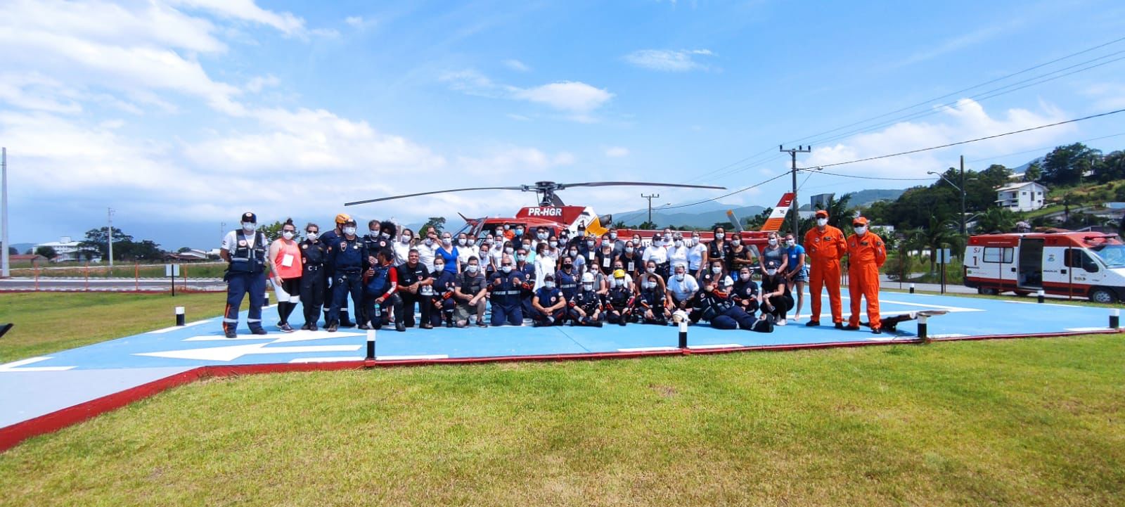 "Grande grupo de profissionais de saúde e resgate posando para foto em heliponto ao lado de helicóptero vermelho do Corpo de Bombeiros com a identificação PR-HGR; ao fundo, ambulâncias e paisagem urbana com céu parcialmente nublado.