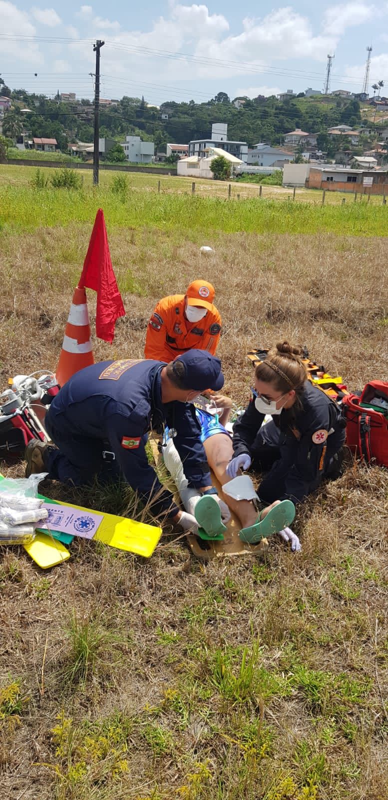 Profissionais de resgate do Corpo de Bombeiros e do SAMU atendem uma vítima de acidente em uma área de campo aberto, utilizando equipamentos de imobilização. A equipe está concentrada no atendimento pré-hospitalar, com materiais espalhados ao redor e um cone com bandeira vermelha sinalizando o local.