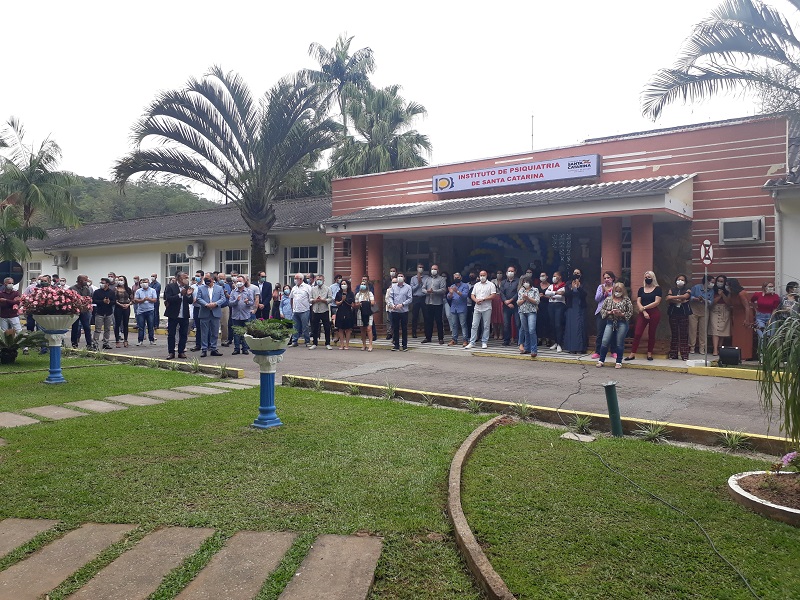 Grupo numeroso de pessoas reunido em frente ao prédio do Instituto de Psiquiatria de Santa Catarina, durante um evento ao ar livre. A fachada do instituto é laranja com detalhes brancos e colunas arredondadas. As pessoas estão organizadas lado a lado, muitas usando máscaras de proteção, e observam atentamente algo fora do quadro. O jardim à frente tem gramado bem cuidado, caminhos de pedra e vasos decorativos com flores.