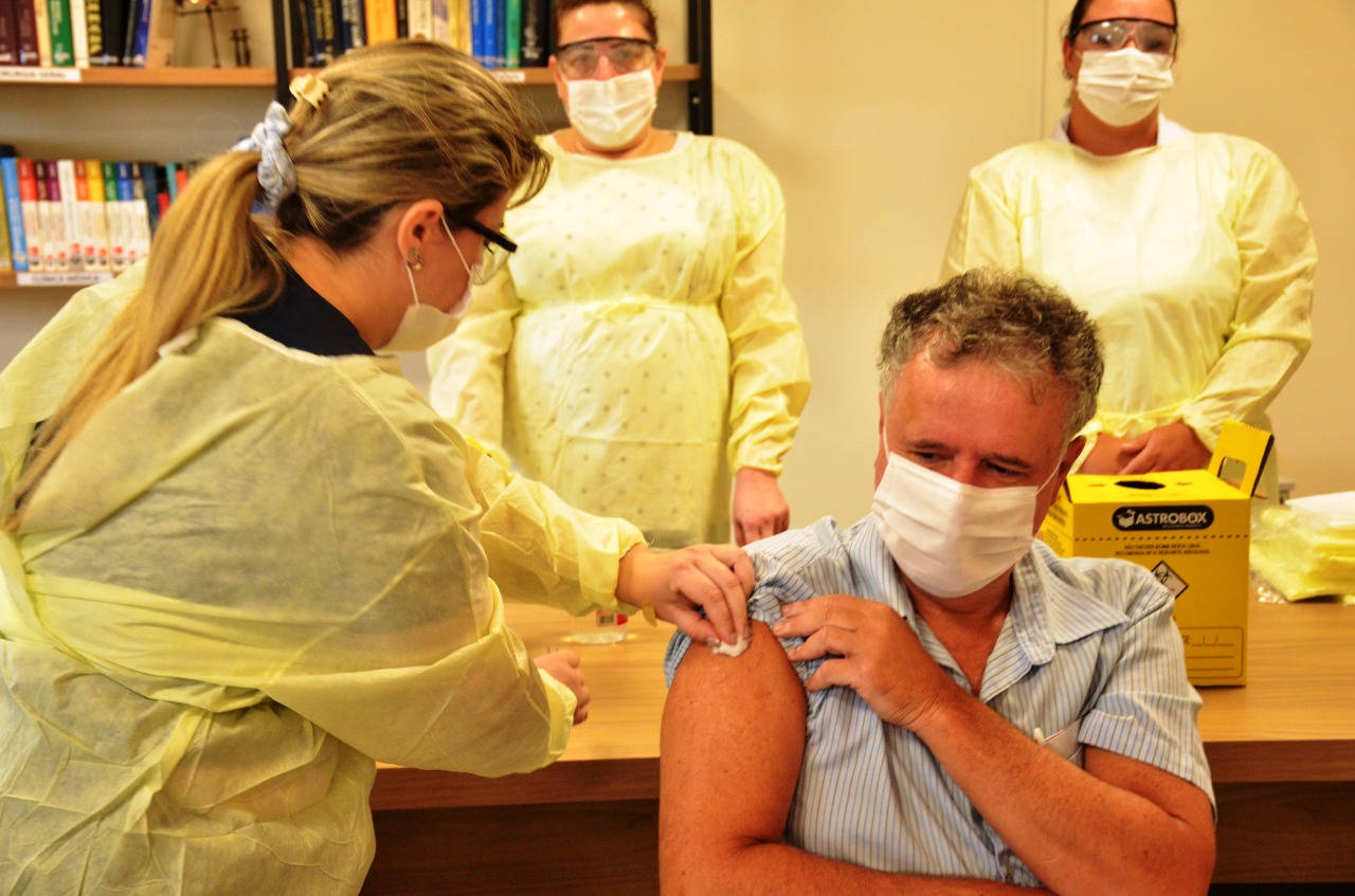 Homem de camisa listrada e máscara facial recebe uma vacina no braço esquerdo, aplicada por uma profissional de saúde usando jaleco amarelo, máscara e óculos de proteção. Ao fundo, três outras profissionais de saúde, também paramentadas, observam a cena.