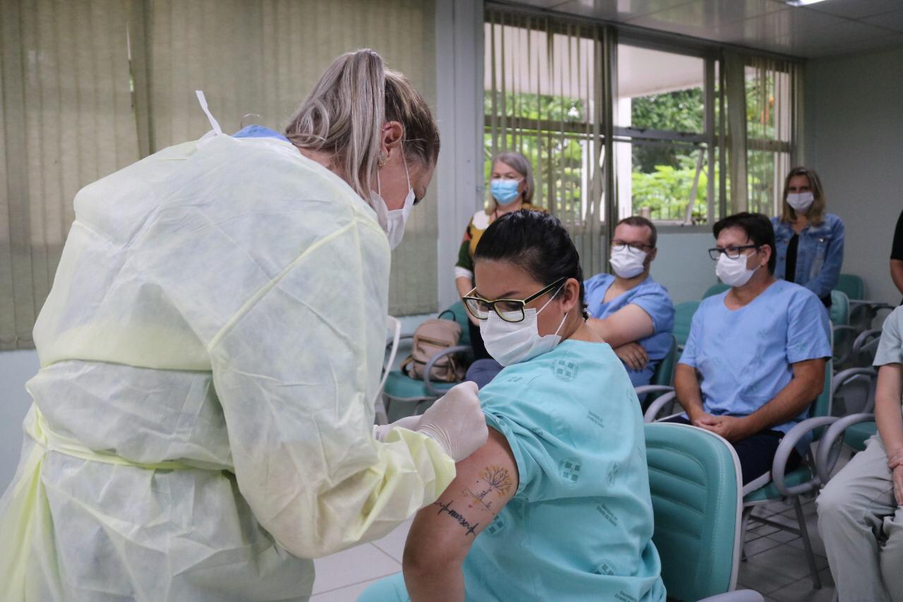Profissional de saúde aplica vacina no braço esquerdo de uma mulher sentada, que veste roupa hospitalar azul e usa máscara. A mulher vacinada tem uma tatuagem visível no braço com desenho de árvore e traçado de eletrocardiograma. 