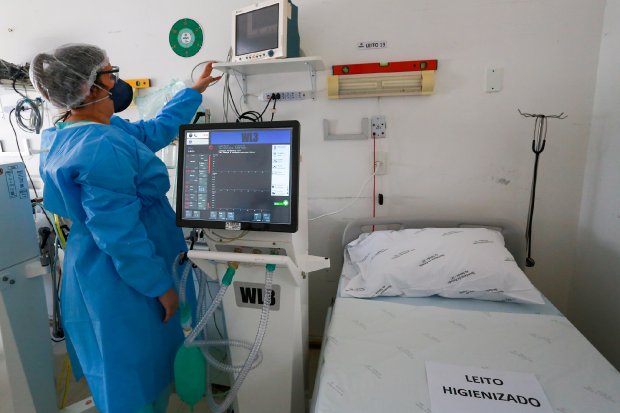 Profissional de saúde com avental azul, touca, máscara e óculos de proteção ajusta um monitor em um leito hospitalar. Ao lado, há um respirador e outro monitor exibindo sinais vitais. A cama hospitalar está vazia, com lençóis brancos e um papel indicando 'LEITO HIGIENIZADO'. O ambiente é limpo e equipado, representando um quarto de UTI pronto para uso.
