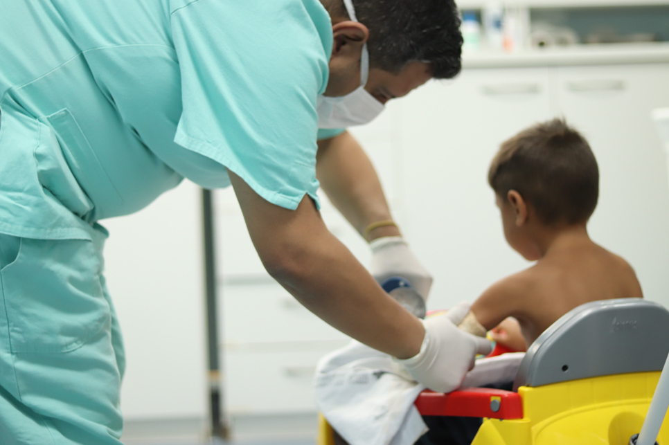 Profissional de saúde usando máscara e luvas realiza atendimento em uma criança sentada em um carrinho infantil dentro de um ambiente hospitalar. A c
