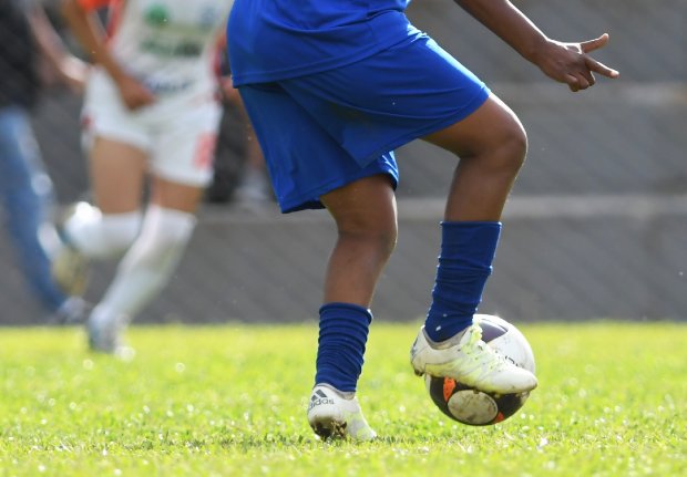 Close nas pernas de uma criança jogando futebol em um campo de grama. Ela veste uniforme azul, incluindo meiões e chuteiras brancas, enquanto domina uma bola de futebol com o pé direito. Ao fundo, desfocado, outro jogador com uniforme branco se aproxima. A cena transmite movimento e concentração durante uma partida.
