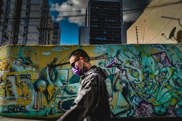 Homem de cabeça raspada e barba curta caminha em frente a um muro coberto por grafites coloridos. Ele usa máscara roxa, fones de ouvido brancos e uma jaqueta escura com capuz. Ao fundo, há edifícios altos, fios elétricos cruzando o céu parcialmente nublado e o contraste entre arte urbana e arquitetura moderna é marcante.