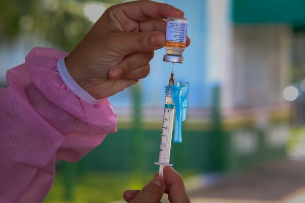 Mão segurando uma seringa sendo preenchida com dose de vacina retirada de um frasco.