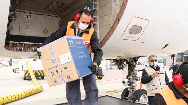 Trabalhador usando colete refletivo, máscara e protetor auricular descarrega uma caixa grande de dentro de um avião no aeroporto. Ao fundo, outras pessoas acompanham a operação, também usando máscaras e coletes, em um ambiente de pista de pouso.