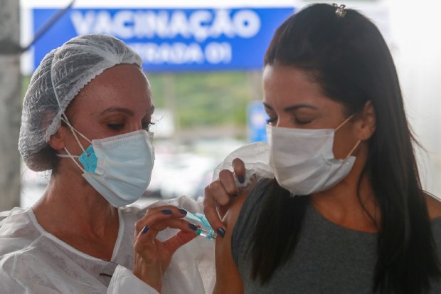 Mulher de roupa branca, máscara branca e touca branca aplicando dose de vacina em mulher de cabelo escuro, máscara branca e blusa cinza