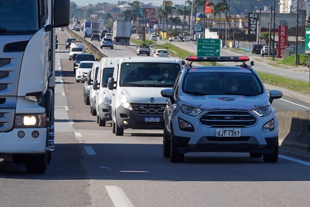 Comboio de veículos trafega por uma rodovia movimentada durante o dia. Na frente, há uma viatura da polícia com luzes de emergência ligadas, seguida por vans brancas. Caminhões e outros carros circulam nas pistas ao lado. Placas verdes de sinalização e comércios são visíveis ao fundo.