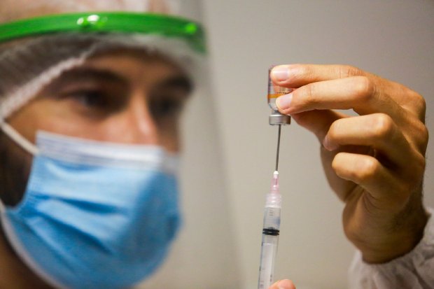 Homem com máscara azul preparando dose de vacina, em uma mão ele segura um frasco, na outra ele insere a seringa no frasco. 