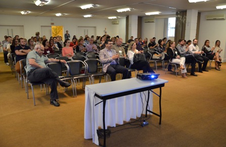 Auditório com dezenas de profissionais da saúde sentados, assistindo atentamente a uma apresentação projetada em um telão (fora do enquadramento).