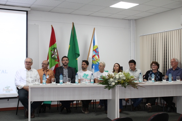 Cena de uma mesa de autoridades composta por nove pessoas, incluindo o Secretário de Estado da Saúde, Dr. Vicente Caropreso, durante a abertura da 2ª Semana de Saúde do Hospital e Maternidade Oase, em Timbó. A mesa está decorada com um arranjo de flores brancas ao centro e garrafas de água à frente de cada participante.